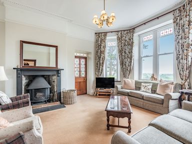A living room with a fireplace a mirror a television sofas armchairs a wooden coffee table and floral curtains at Pensarn Hall in Llanbedr near Harlech