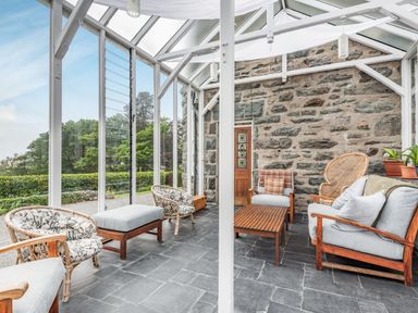 A conservatory with wooden chairs, a coffee table, and a stone wall at Pensarn Hall in Llanbedr near Harlech