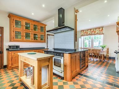 A kitchen with a wooden island, stove, wooden cabinets, a dining table by a window with patterned curtains at Pensarn Hall Llanbedr near Harlech