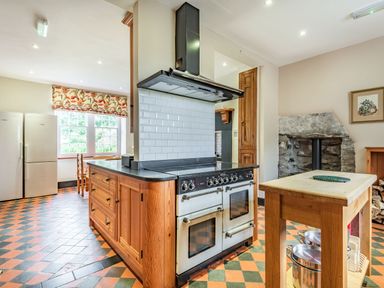 A kitchen with a wooden island and a double oven stove a small wooden table with pots and pans two refrigerators a window with floral curtains and a stone fireplace at Pensarn Hall in Llanbedr near Harlech