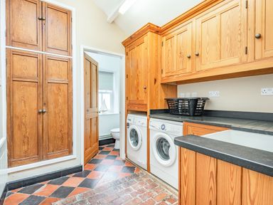 A laundry room with wooden cabinets a washing machine a dryer a black laundry basket and a door leading to a bathroom at Pensarn Hall in Llanbedr near Harlech