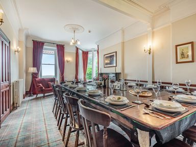 A dining room with a long wooden table set with plates glasses and cutlery surrounded by wooden chairs with red curtains and an armchair by windows at Pensarn Hall Llanbedr near Harlech