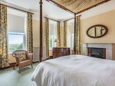 A bedroom with a four-poster bed a wicker chair floral curtains and a fireplace at Pensarn Hall in Llanbedr near Harlech