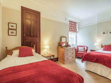 A bedroom with two single beds with red blankets a wooden dresser a red armchair and a window with red checked blinds at Pensarn Hall in Llanbedr near Harlech