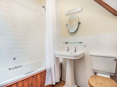 A bathroom with a bathtub wooden panel white shower curtain pedestal sink with two taps round mirror and toilet with wooden lid at Pensarn Hall in Llanbedr near Harlech