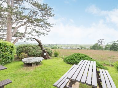 An outdoor garden area with a wooden picnic table stone table large tree bushes and fields in the background at Pensarn Hall in Llanbedr near Harlech