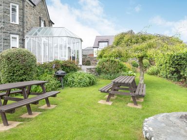 A garden with two wooden picnic tables a barbecue grill and a glass conservatory attached to a stone house at Pensarn Hall in Llanbedr near Harlech