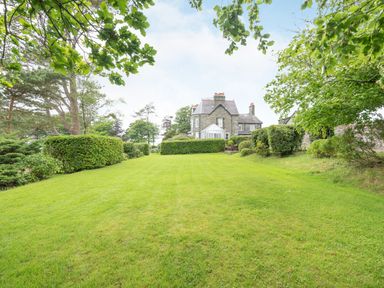 A large lawn with trimmed hedges and trees surrounding a stone house with a conservatory at Pensarn Hall in Llanbedr near Harlech