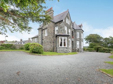 A stone house with multiple windows and a gravel driveway surrounded by bushes and trees at Pensarn Hall in Llanbedr near Harlech