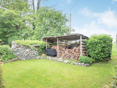 A garden area with stacked firewood in a wooden shelter and a stone wall with greenery at Pensarn Hall in Llanbedr near Harlech