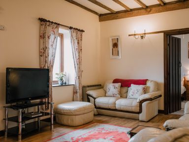 A living room with a cream sofa with floral cushions a matching ottoman a television on a stand and floral curtains at Stargazer's Field House Hardwicke near Hay-on-Wye