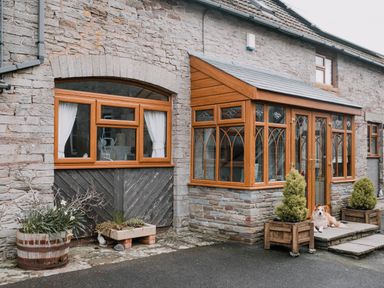 The exterior of a stone house with wooden framed windows a small porch with glass walls two potted plants and a dog sitting on the porch at Stargazer's Field House in Hardwicke near Hay-on-Wye