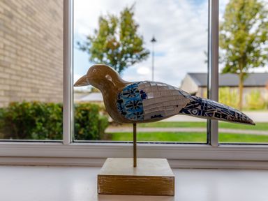 A bird sculpture with a mosaic pattern on a stand on a windowsill with trees outside at Perriwinkle The Bay near Filey