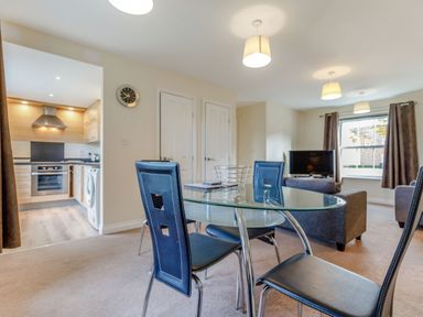 A dining area with a glass table and four chairs next to a living room with a sofa and a TV near a window at Perriwinkle The Bay near Filey