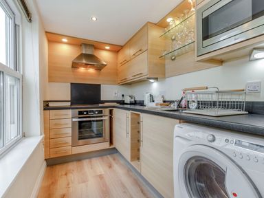A kitchen with wooden cabinets a stove a washing machine a microwave and glass shelves at Perriwinkle The Bay near Filey