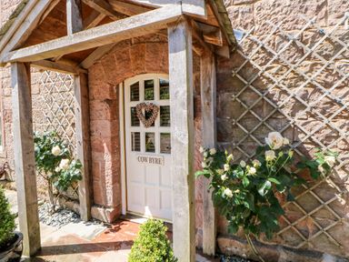 An entrance door with a wooden porch and climbing plants on trellis on the stone wall at The Cow Byre in Barras near Kirkby Stephen