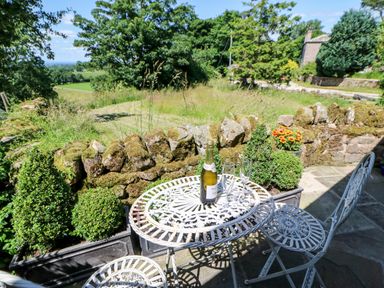 An outdoor patio with a white metal table and chairs a bottle and two glasses on the table plants and a stone wall in the background at The Cow Byre in Barras near Kirkby Stephen