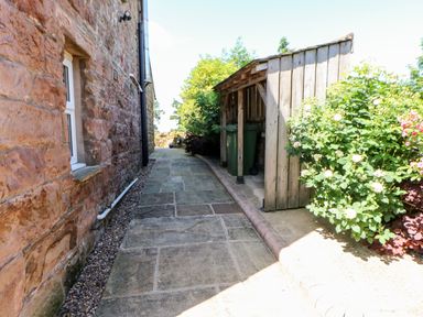 A stone pathway beside a brick wall with a wooden shed and green bins next to flowering shrubs at The Cow Byre in Barras near Kirkby Stephen