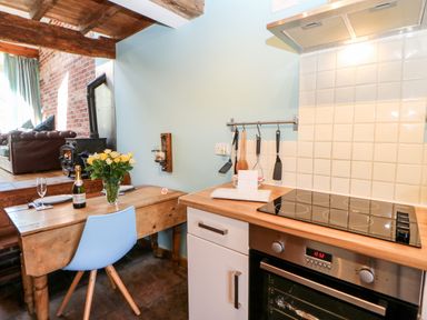 A kitchen with an electric stove oven and utensils hanging on a wall next to a wooden table with a blue chair vase of yellow flowers and bottle of wine at The Cow Byre Barras near Kirkby Stephen