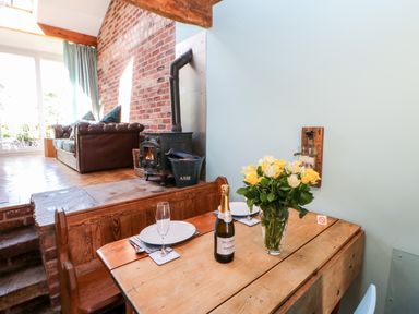 A dining table with flowers and a bottle next to a living area with a sofa and wood stove at The Cow Byre in Barras near Kirkby Stephen