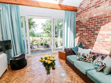A sitting room with a glass table and flowers next to a leather sofa and sliding glass doors at The Cow Byre in Barras near Kirkby Stephen