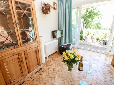 A room with a wooden cabinet flowers and champagne on a clear table and a sliding glass door opening to a patio at The Cow Byre in Barras near Kirkby Stephen