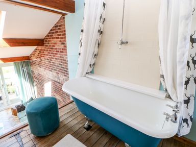 A blue clawfoot bathtub with deer print curtains in a bathroom overlooking a living area with exposed brick walls at The Cow Byre Barras near Kirkby Stephen