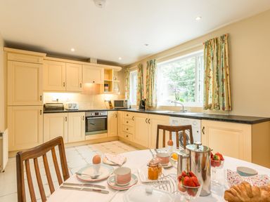 A kitchen with yellow cabinets a dining table set with breakfast items and a window with floral curtains at Stephen's Cottage in Alnmouth