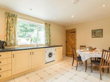 A kitchen with cabinets a washing machine a window with floral curtains and a dining table set for breakfast at Stephens Cottage in Alnmouth
