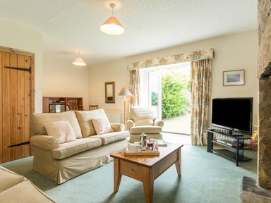 A living room with sofas a wooden coffee table a television and an open door with floral curtains at Stephens Cottage in Alnmouth