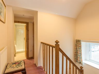 A staircase landing with wooden banister a window with floral curtains a bench with embroidered cushion and an open door to a bathroom at Stephens Cottage in Alnmouth