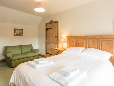A bedroom with a wooden bed headboard white bed linens folded towels a green sofa a wooden door and a bedside table lamp at Stephens Cottage in Alnmouth