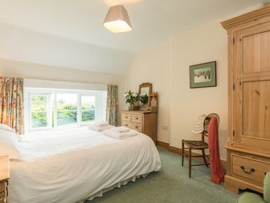 A bedroom with a bed and white bedding near a window with floral curtains a wooden dresser with a mirror a wooden chair with a hat and scarf and a wooden wardrobe at Stephens Cottage in Alnmouth