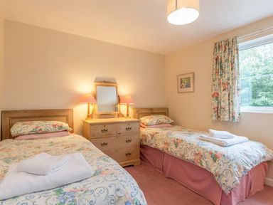 A bedroom with two single beds with floral bedding a wooden dresser with a mirror and two lamps a window with floral curtains at Stephens Cottage in Alnmouth