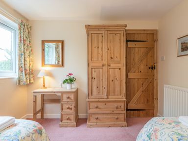 A bedroom with a wooden wardrobe a desk with a lamp and a plant floral curtains and a wooden door at Stephen's Cottage in Alnmouth