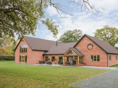 An outdoor view of a brick building with a patio and chairs surrounded by grass and trees at Oak Lodge in Beaconsfield Holiday Park