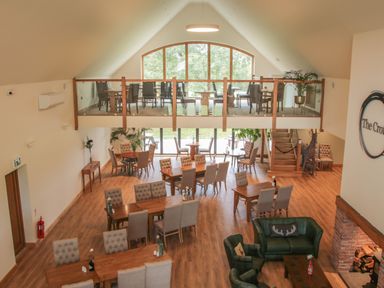 A dining area with wooden tables and upholstered chairs and a balcony with additional seating at Oak Lodge in Beaconsfield Holiday Park
