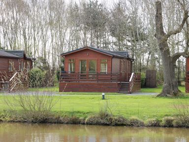 A wooden lodge with a deck and outdoor furniture near a pond with trees in the background at Oak Lodge in Beaconsfield Holiday Park