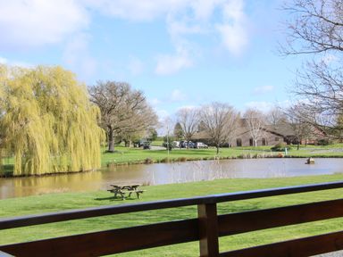A pond with trees and picnic benches in a park with buildings and parked cars at Oak Lodge Beaconsfield Holiday Park