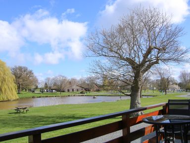 A view of a lake a tree and picnic table from a wooden deck with a glass table and chairs at Oak Lodge in Beaconsfield Holiday Park