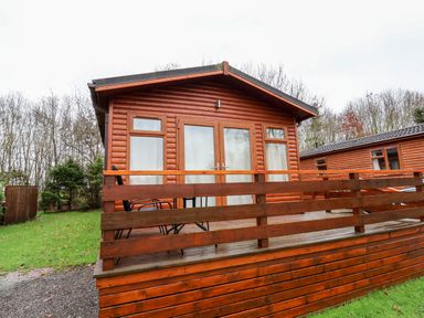 A wooden lodge with a deck railing and outdoor chairs at Oak Lodge in Beaconsfield Holiday Park
