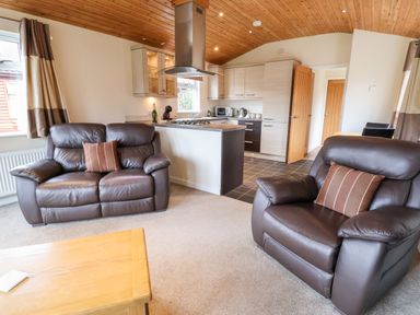 A living room with two brown leather armchairs a kitchen with light wood cabinets and a wooden ceiling at Oak Lodge in Beaconsfield Holiday Park