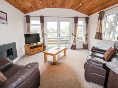 A living room with brown leather sofas a wooden coffee table a wooden TV stand with a television and glass doors with curtains at Oak Lodge Beaconsfield Holiday Park