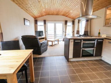 An open plan kitchen and living area with wooden dining table black chairs black recliner and cooking island with oven at Oak Lodge in Beaconsfield Holiday Park