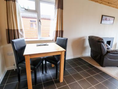 A dining area with a wooden table and two black chairs next to a window and a living area with a brown leather sofa at Oak Lodge in Beaconsfield Holiday Park