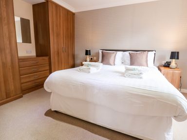 A bedroom with a large bed with white bedding and brown cushions bedside tables with lamps and wooden wardrobe Oak Lodge Beaconsfield Holiday Park