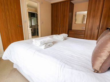 A bedroom with a bed with white linens and towels two wooden wardrobes a chest of drawers and a door leading to a bathroom at Oak Lodge Beaconsfield Holiday Park