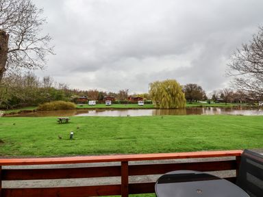 A patio with a round table overlooking a grassy area with picnic benches and a pond with caravans and trees at Oak Lodge in Beaconsfield Holiday Park