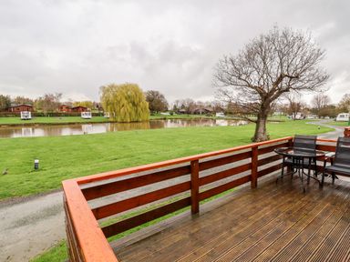 A wooden deck with a round glass table and two chairs overlooking a grassy area with a pond and caravans at Oak Lodge in Beaconsfield Holiday Park