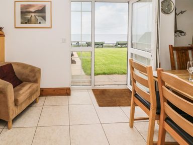 A room with a brown armchair a wooden dining table and chairs and a glass door opening to a grassy area and sea view at On The Watersedge in Beadnell